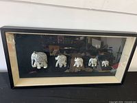 Front view of framed shadow box showing five silver-tone elephant figurines against black background and cream mat