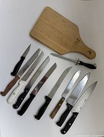 Full group shot of nine knives, honing steel and wooden cutting board