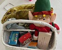 Top view of basket showing gold tree skirt, elf plush, boxed alarm clock, red package, sleep mask box