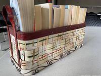 Side view of woven basket holding paperback books, showing wooden rim, handle, painted stripe design, metal scroll base, tops of books visible