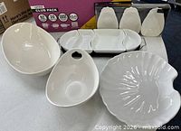 All white ceramic serving pieces laid out including shell platter, bowls, divided tray on wooden base, packaged pineapple dishes