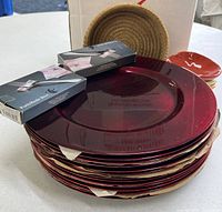 Stack of red charger plates with two Fortessa boxes of utensil rests, one red pickle dish, and woven bread basket visible