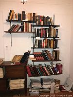 Full view of seven wooden wall-mounted shelves filled with vintage books, wooden side cabinet with more books and papers below