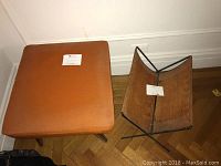 Mid-Century Modern padded orange ottoman next to vintage folding suede magazine rack on wooden floor.