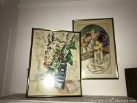 Photo showing two framed vintage watercolor still life paintings. The left painting shows a blue square vase with flowers and an open book beside it. The right painting features a curved composition of flowers in a vase.