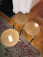 Top view of three round wooden top snack tables with metal frames, arranged side by side on wood floor and patterned rug.