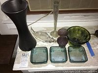 Full view of all glass items on a wooden surface including tall black vase, clear fish dish, slender clear vase, purple vase, green bowl and three square aqua tinted glass plates.