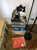 Photo showing top and side view of wheeled plastic storage unit with four clear drawers; various office supplies visible on top including pens, scissors, hole puncher, Epson 126 ink cartridges, and boxes of labels and paper next to unit.