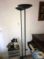 Full view of two black metal floor lamps with bowl-shaped upward facing shades next to a piano and drawer unit.