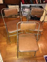 Three vintage Samsonite folding chairs grouped together, showing metal frame and brown vinyl cushions, placed on wooden parquet floor.