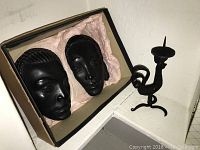 Two black painted women's wall masks in the original box with tissue paper and a hand forged metal bird shaped candle holder on the side.