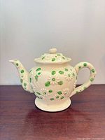 Left side view of cream porcelain teapot with green leaves and white floral embossing