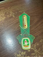 Full view of green and gold metal wall thermometer showing scale and commemorative badge
