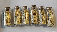 Six small clear glass vials with cork stoppers filled with gold leaf flakes displayed in a row