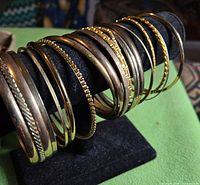 Group of approximately 18 gold-tone bangles on a display rod showing range of widths and textures