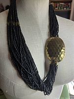 Necklace on bust showing multiple black seed bead strands and hammered gold-tone oval accent