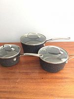 Three black speckled saucepans with glass lids displayed on tabletop