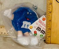 Front view of sealed blue M&M plush ornament beside ruler showing size