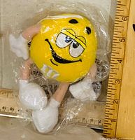 Front view of yellow M&M plush ornament in sealed bag next to ruler for scale