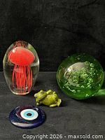 Group view of red jellyfish glass paperweight, green bubble glass paperweight, blue evil-eye disk, and green ceramic frog figurine