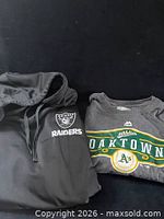 Front view of black Raiders hoodie beside charcoal Athletics T-shirt