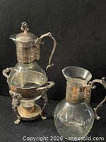Coffee/tea server in ornate stand next to separate glass pitcher/carafe