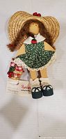 Front view of wooden Punkins doll wearing green floral dress, straw hat, holding basket with tag