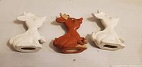 Front view of three seated ceramic reindeer, two white bisque, one brown painted