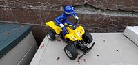 Oblique view of yellow quad bike toy with blue rider on outdoor surface