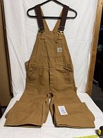 Front view of brown Carhartt bib overalls laid flat showing full length and suspenders