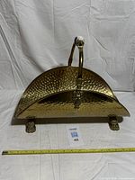 Full view showing hammered brass log holder, handle, lion face, paw feet, measuring tape for scale