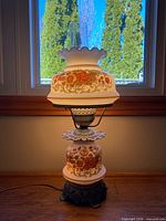 Front view of lit double light floral glass table lamp showing shade, chimney, floral base, and cord
