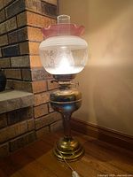 Full view lit brass converted oil lamp with etched globe and pink ruffled shade