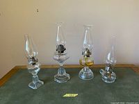 All four clear glass oil lamps lined up showing shape, bases, burners and chimneys