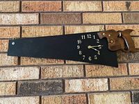 Black painted hand saw wall clock with wood handle mounted on brick wall