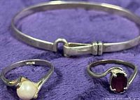 Top-down view of bangle, pearl ring, garnet ring on purple fabric