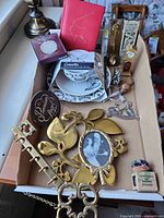 Overview of box showing brass decor, china tea set, figurines, plaque, frame, key holder and other small items