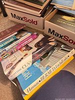 Stack of reference and travel books with visible titles including Scottish Surnames, Scotland guides, Bermuda and Venice plus filled boxes of additional books