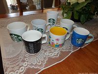 Seven Starbucks mugs on lace tablecloth