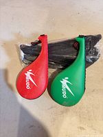 Red and green Mudo kicking paddles beside sealed package of shin guards