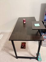 Angled view of desk surface, showing dark brown top, black metal frame, small elevated shelf, can for scale