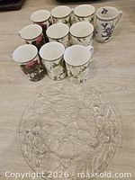 Ten floral bone china mugs and clear glass serving plate on floor