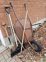 Wheelbarrow with assorted garden tools leaning against brick wall