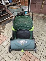 Front view of manual lawn sweeper showing Sweep It logo, wheels, brush housing and bag