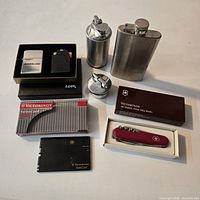 Group shot of all items: SwissCard in box, Swiss Army knife boxed, Zippo box open, two Ronson lighters, hip flask