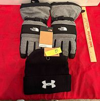 Pair of The North Face Montana Ski Gloves and Under Armour beanie on red surface with ruler for scale