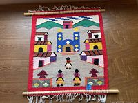 Full view of the hand-woven wall hanging showing entire village scene, red border, dowels and fringe