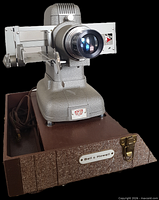 Front angled view of Bell & Howell projector on carrying case base showing lens, slide gate arm, power cord, and brand plate