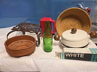 Group view of all items including cast iron stand, terracotta saucer, red pitcher, green bottle, white fondue pot, wicker basket, paint tube