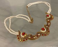 Necklace laid flat showing triple pearl strands, metal centerpiece, red stones, clasp and extender